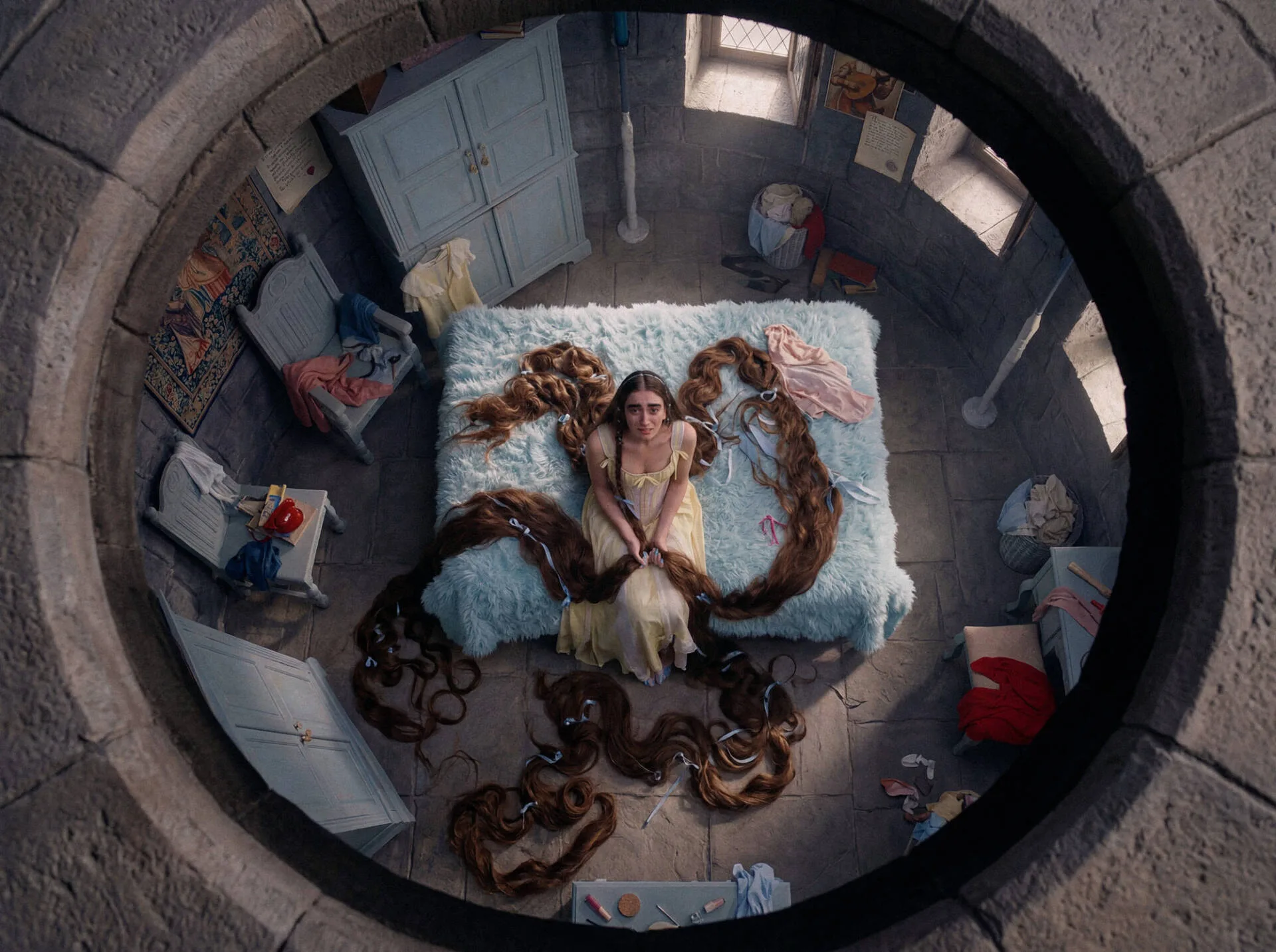 Viewed through a circular stone opening, Rapunzel sits on a blue bed with long brown hair coiled across the tower room