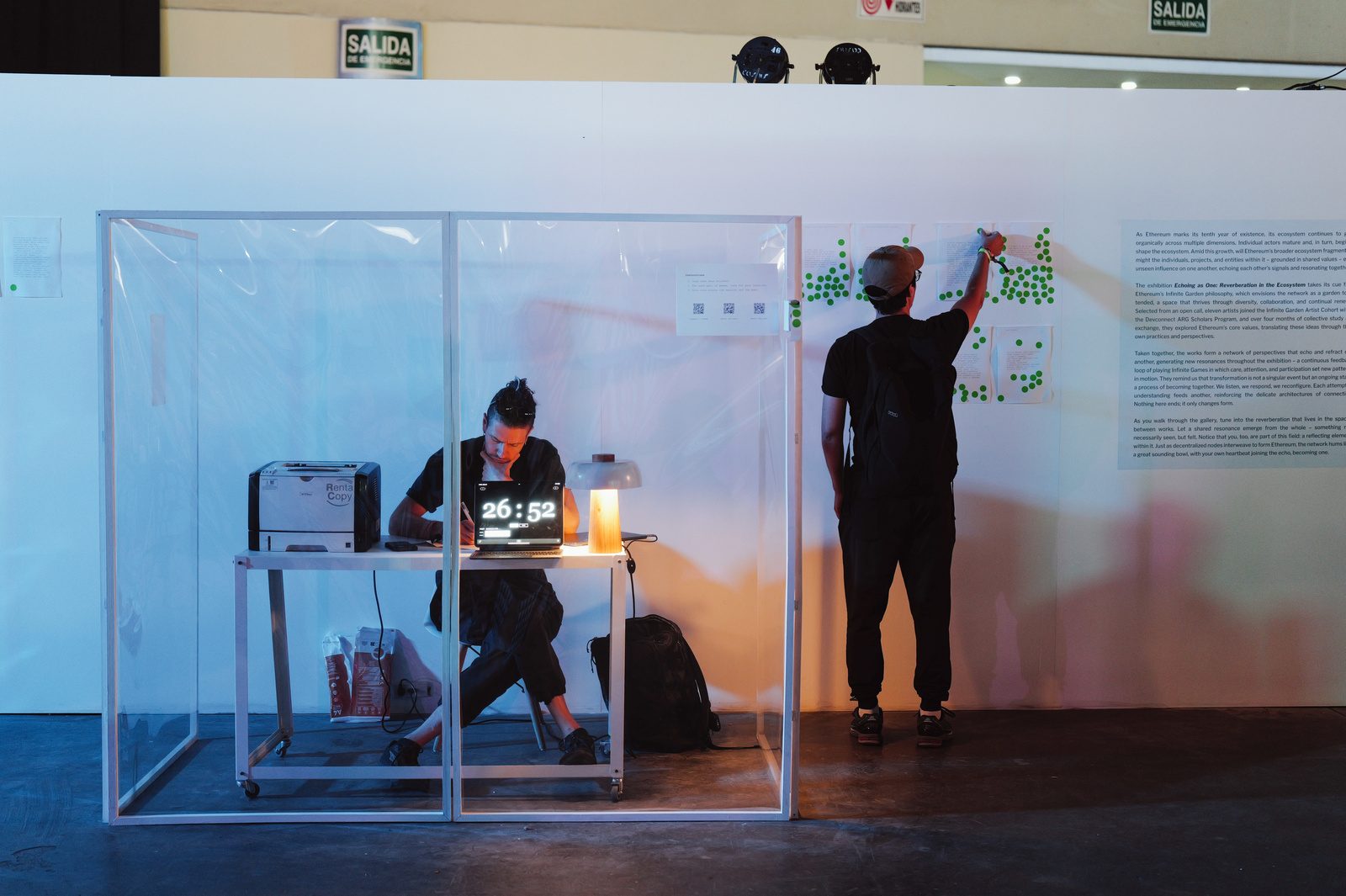 A poet writes inside a transparent cube while a visitor adds a green voting sticker to poems on the wall