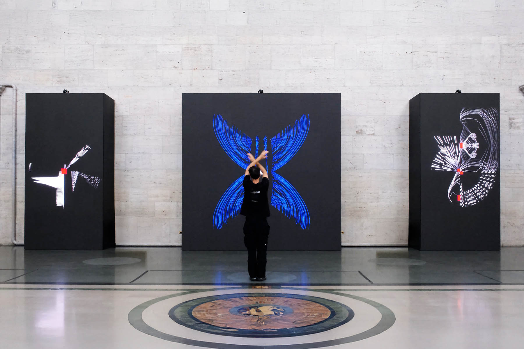 A visitor raises their hands before three black monoliths showing blue and white movement patterns